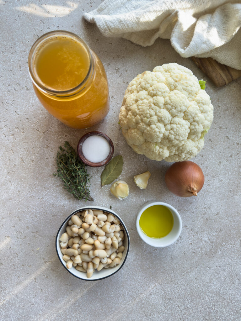 whole head of cauliflower, vegetable stock, white beans, onion and a variety of herbs and spices for a white bean soup on the counter.
